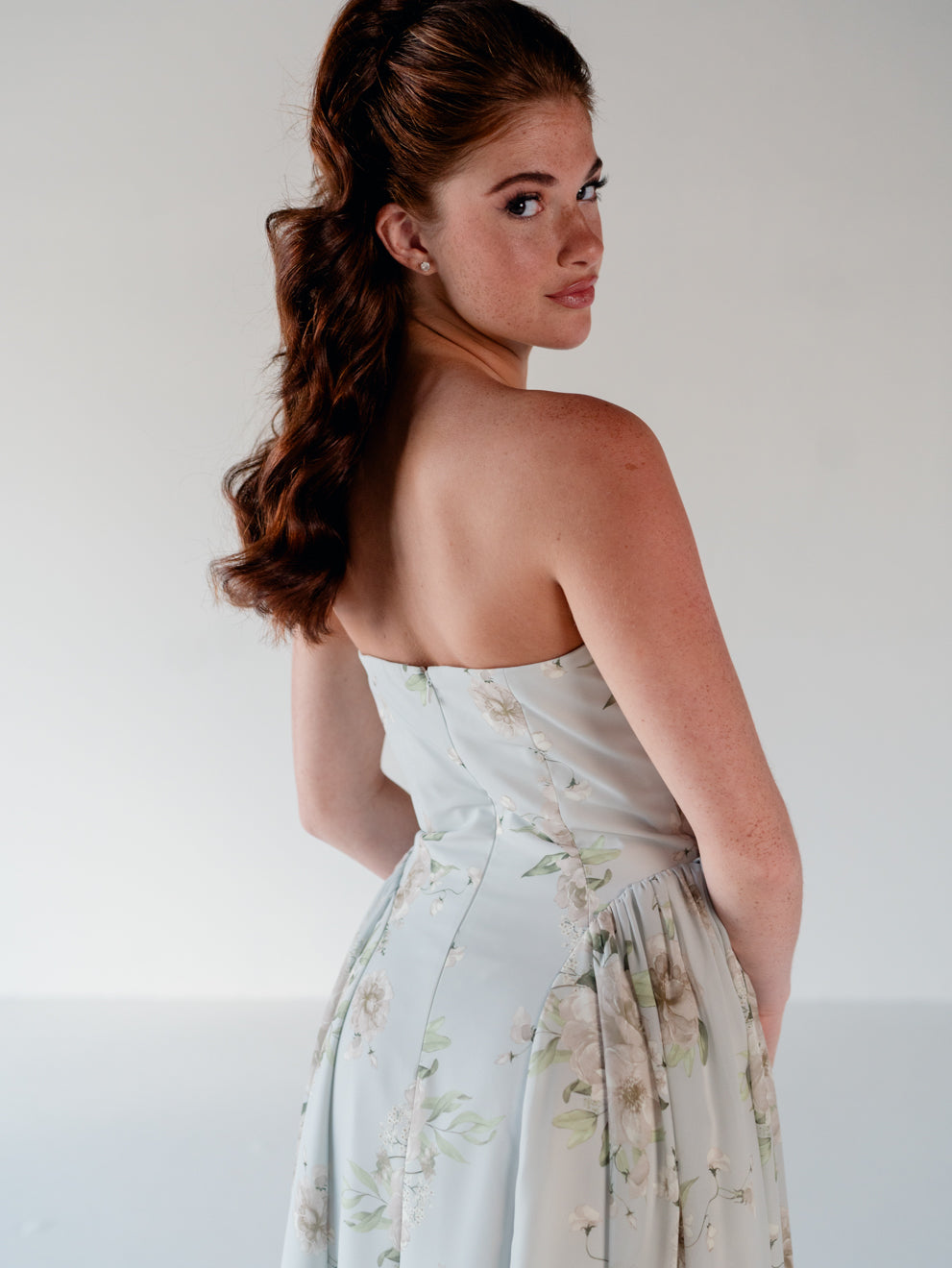 A woman with long auburn hair is shown from behind wearing a strapless light blue-green floral dress against a plain light gray studio background. / Draped neckline gown with gathered side detail.