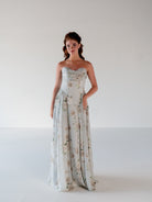 A woman stands in a studio wearing a strapless floor-length gown with a fitted bodice and a flowing floral skirt. The dress is a very light icy blue or pale gray base with soft beige and muted green floral print. / Draped neckline gown with gathered side detail.
