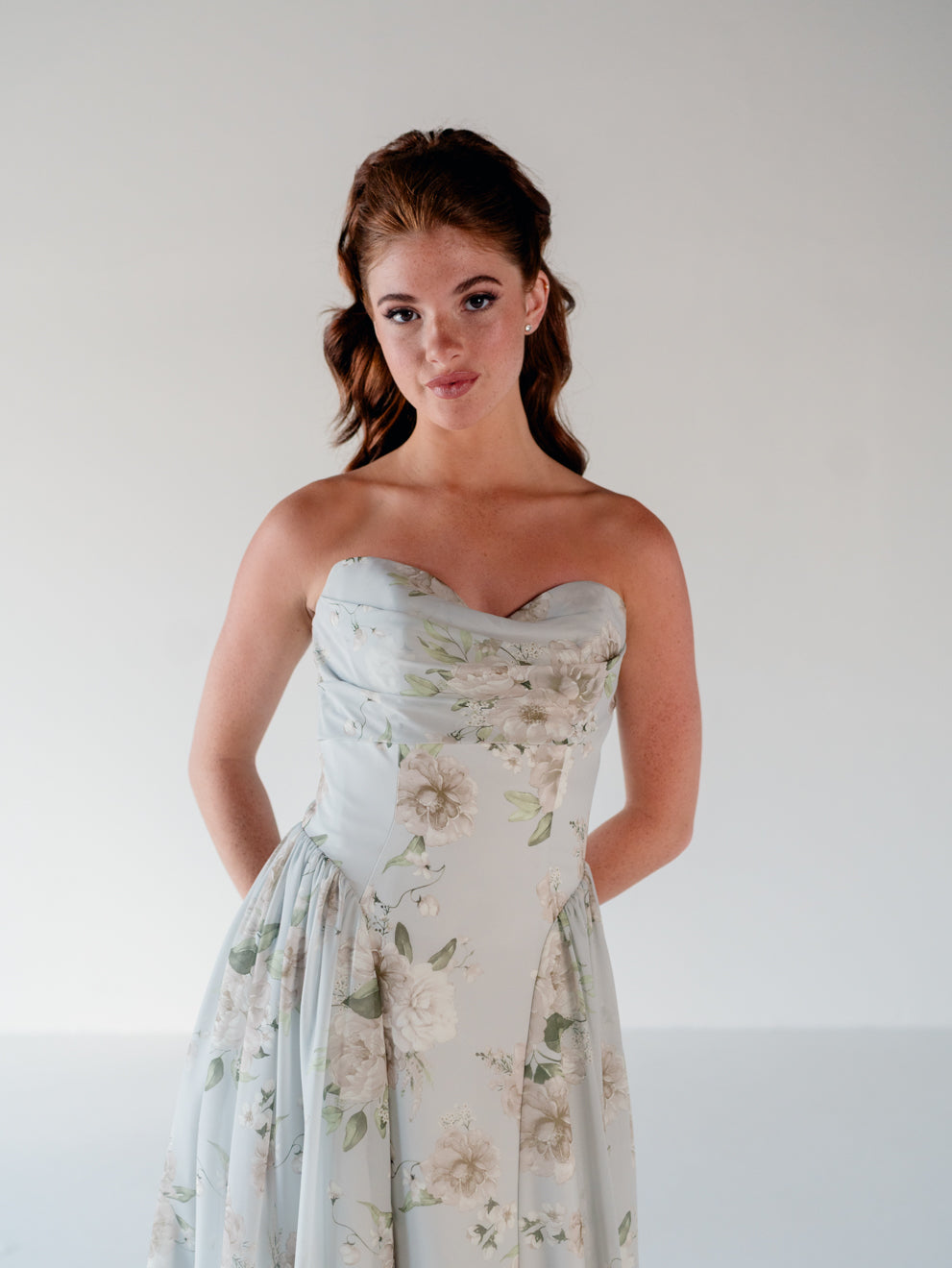 A woman wearing a strapless sweetheart-neck dress with a light blue base and a soft beige and green floral print, posed against a plain light gray studio background. / Draped neckline gown with gathered side detail.