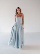 A woman modeling a strapless light blue floor-length gown with a gathered bodice and flowing skirt, standing against a plain white studio background. / Draped neckline gown with gathered side detail.
