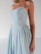 A woman wearing a strapless light blue dress with a structured sweetheart bodice and a flowing skirt, shown against a plain light gray studio background. / Draped neckline gown with gathered side detail.