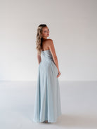 A woman modeling a strapless light blue maxi dress, photographed from the back in a minimalist studio setting with a white background. / Draped neckline gown with gathered side detail.
