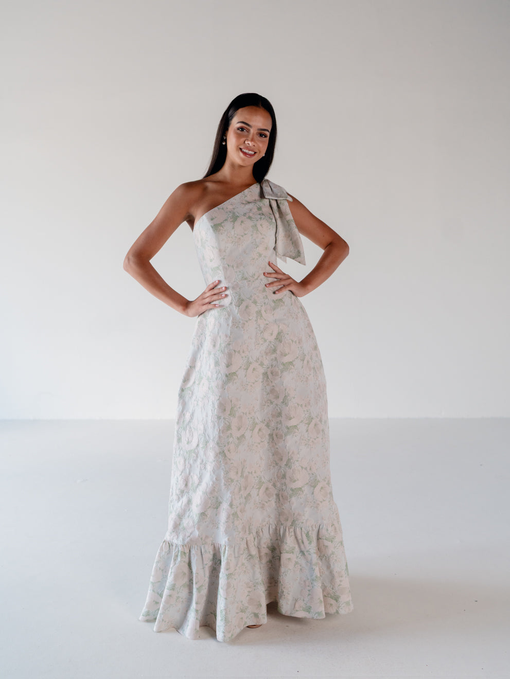 A woman modeling a one-shoulder maxi dress with a textured, floral jacquard pattern in soft beige, white, and muted sage green. The dress features a bow detail on the shoulder and a ruffled hem, set against a plain white studio background. / One shoulder gown with bow detail and ruffle at the skirt.
