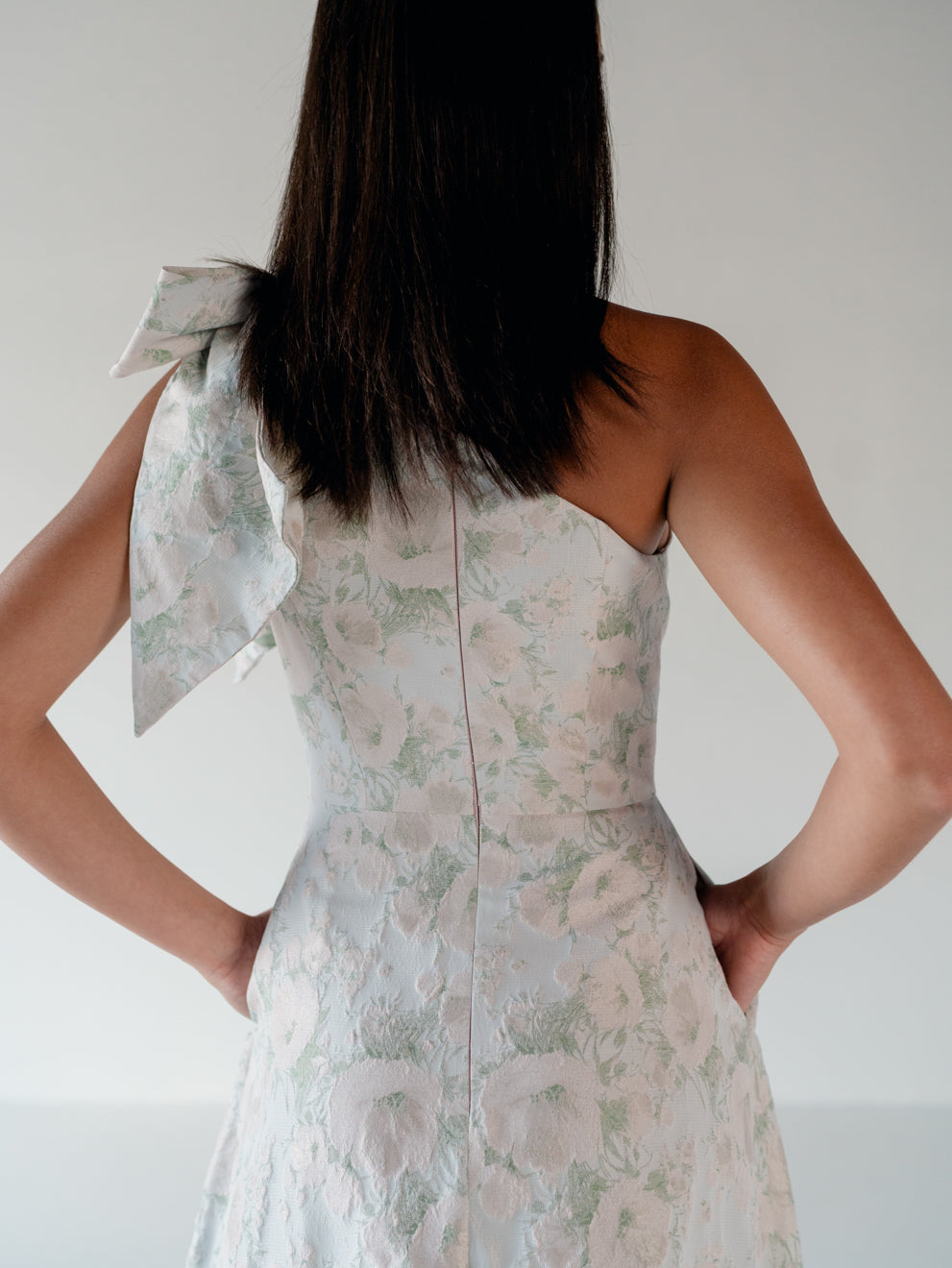 A woman shown from the back wearing a sleeveless floral dress with a large bow detail on one shoulder, set against a plain light gray background. / One shoulder gown with bow detail and ruffle at the skirt.