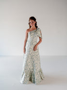 A woman modeling a one-shoulder floor-length gown with a delicate floral print in sage green and cream, set against a plain white studio background. / One shoulder gown with bow detail and ruffle at the skirt.