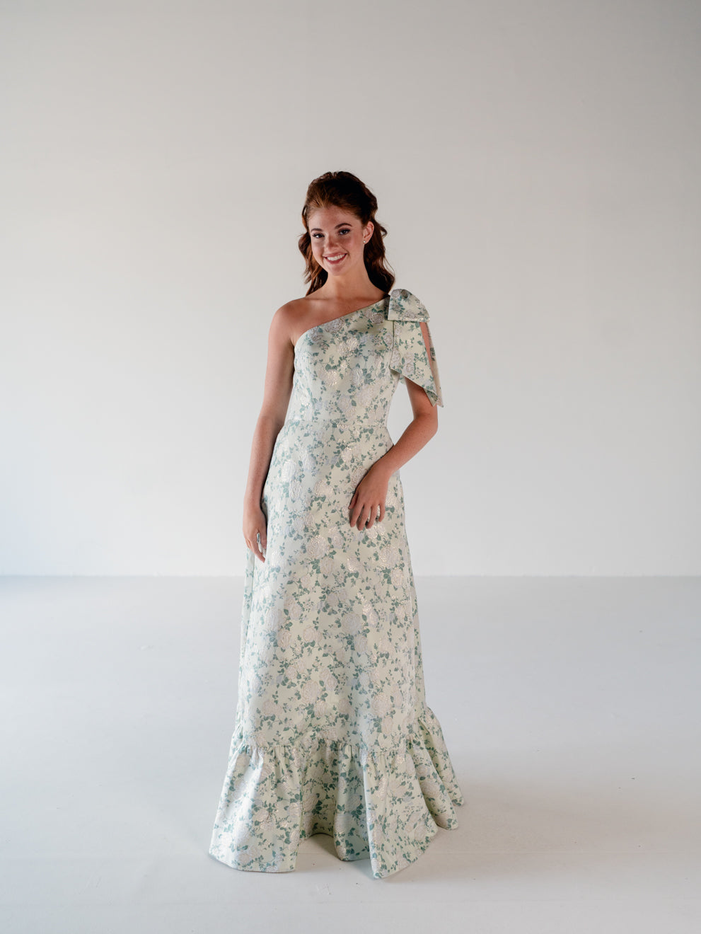 A woman modeling a one-shoulder floor-length gown with a delicate floral print in sage green and cream, set against a plain white studio background. / One shoulder gown with bow detail and ruffle at the skirt.