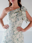 A woman wearing a one-shoulder formal dress with a large bow detail on the shoulder, featuring a light beige base with a green floral pattern and metallic rose-gold floral highlights. / One shoulder gown with bow detail and ruffle at the skirt.