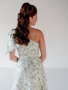 A woman shown from the back wearing a one-shoulder midi dress with a floral pattern in sage green and cream, featuring a large bow detail on the shoulder. / One shoulder gown with bow detail and ruffle at the skirt.