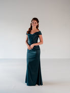 A woman modeling a floor-length off-the-shoulder gown in a deep teal satin-like fabric, standing against a plain light gray studio background. / Off the shoulder, stretch matte charmeuse gown with asymmetrical draping.