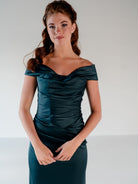A woman modeling an off-the-shoulder dark teal satin dress with ruched detailing on a light gray studio background. / Off the shoulder, stretch matte charmeuse gown with asymmetrical draping.