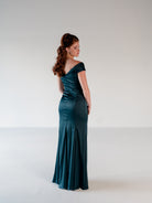 A woman stands in profile wearing a floor-length satin evening gown in a deep teal green color, photographed against a seamless light gray studio background. / Off the shoulder, stretch matte charmeuse gown with asymmetrical draping.