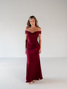 A woman modeling a floor-length off-the-shoulder gown in deep burgundy with a ruched bodice and satin-like sheen, standing against a plain light gray studio background. / Off the shoulder, stretch matte charmeuse gown with asymmetrical draping.