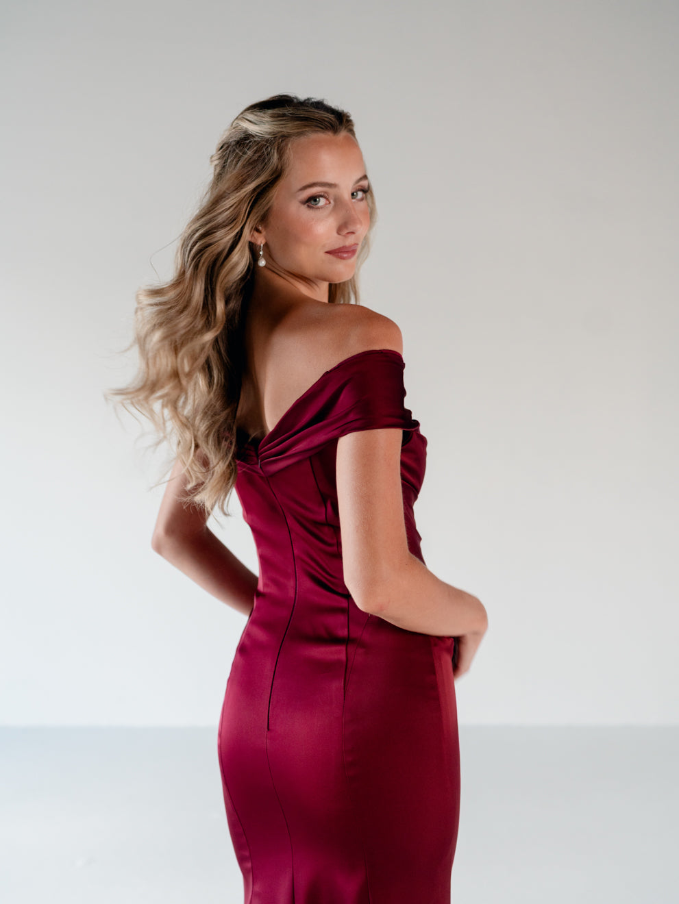 A woman modeling an off-the-shoulder, satin-like burgundy evening gown against a plain light gray studio background. / Off the shoulder, stretch matte charmeuse gown with asymmetrical draping.