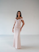 A woman modeling a floor-length off-the-shoulder gown in a pale blush pink color, standing against a neutral light gray studio background. / Off the shoulder, stretch matte charmeuse gown with asymmetrical draping.