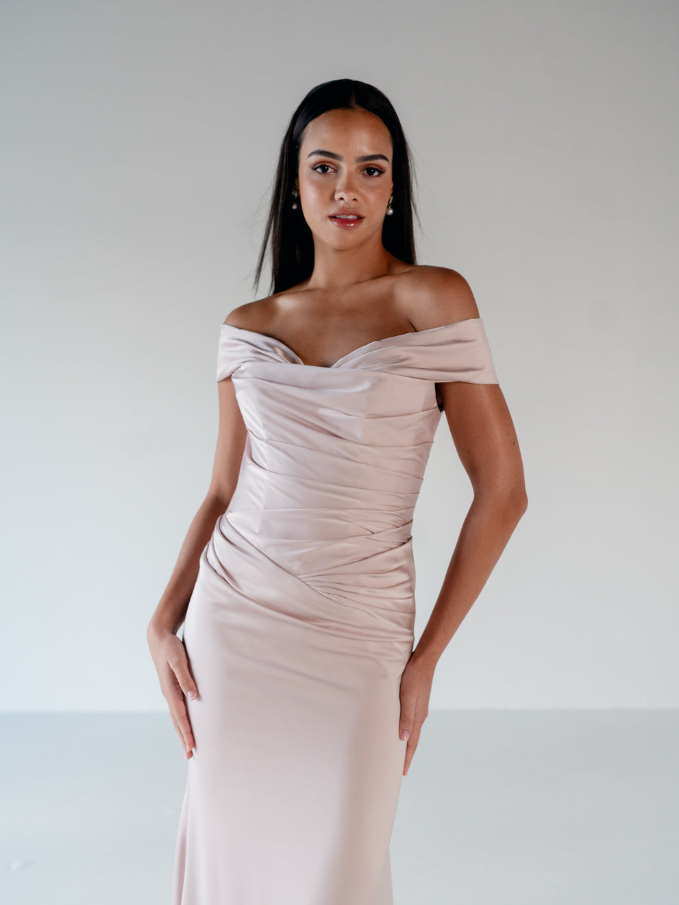A woman modeling an off-the-shoulder, ruched floor-length gown in a soft blush pink color, set against a plain light gray studio background. / Off the shoulder, stretch matte charmeuse gown with asymmetrical draping.