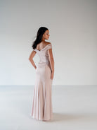 A woman modeling a floor-length off-the-shoulder gown in a pale blush pink color, photographed from the back in a minimalist studio setting with a light gray background. / Off the shoulder, stretch matte charmeuse gown with asymmetrical draping.