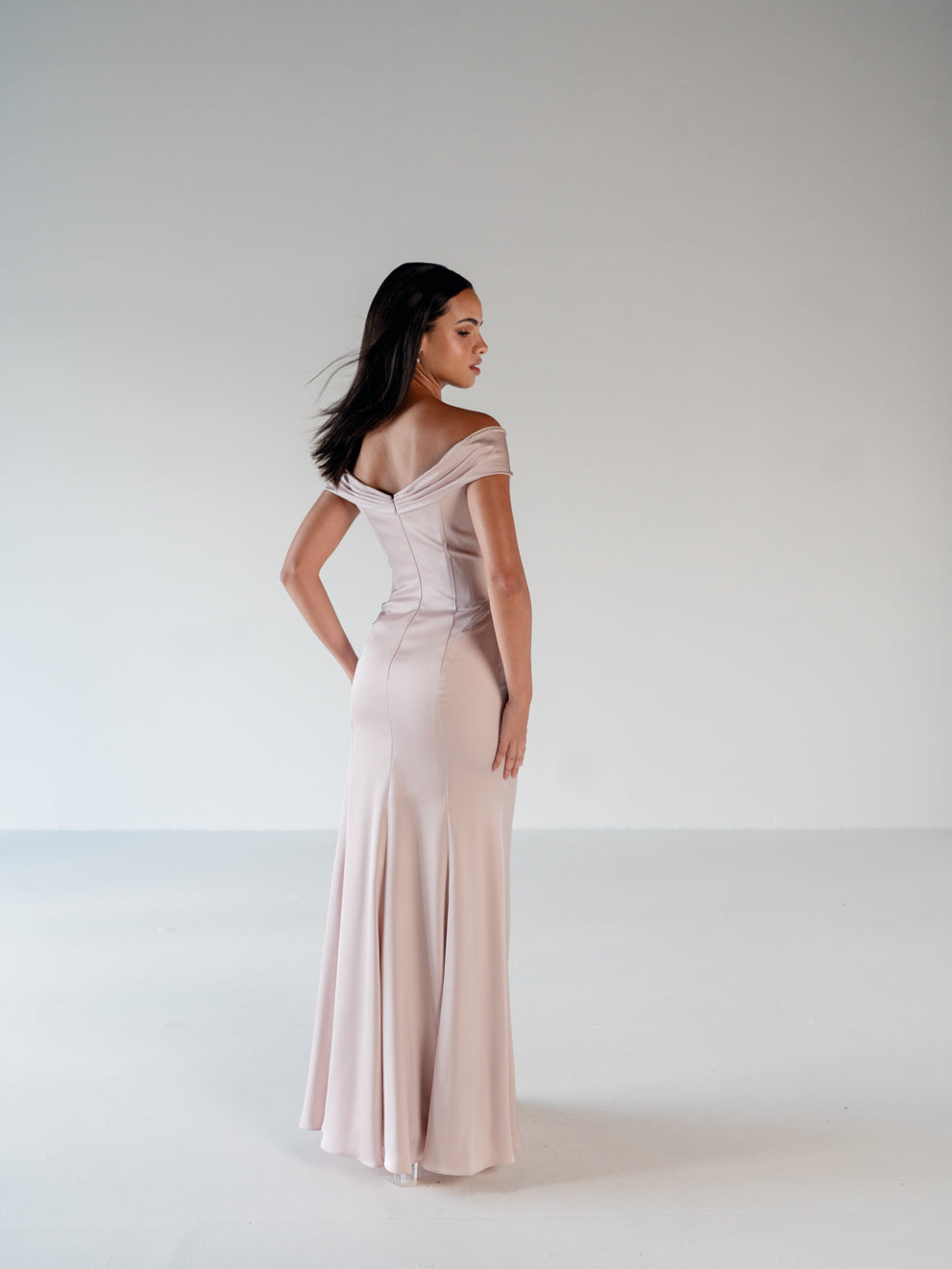 A woman modeling a floor-length off-the-shoulder gown in a pale blush pink color, photographed from the back in a minimalist studio setting with a light gray background. / Off the shoulder, stretch matte charmeuse gown with asymmetrical draping.