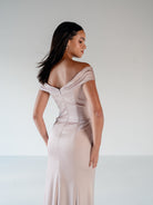 A woman wearing a floor-length off-the-shoulder gown in a pale blush pink satin, photographed from the back against a neutral light gray studio background. / Off the shoulder, stretch matte charmeuse gown with asymmetrical draping.