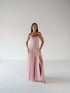 A woman modeling a floor-length, strapless dusty pink gown with a draped bodice, flowing skirt, and high front slit, standing against a plain light gray studio background. / Strapless draped bodice with asymmetrical placed skirt and lace up back.