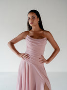 A woman modeling a strapless dusty pink dress with a draped, ruched bodice and a high side slit, set against a plain light gray studio background. / Strapless draped bodice with asymmetrical placed skirt and lace up back.