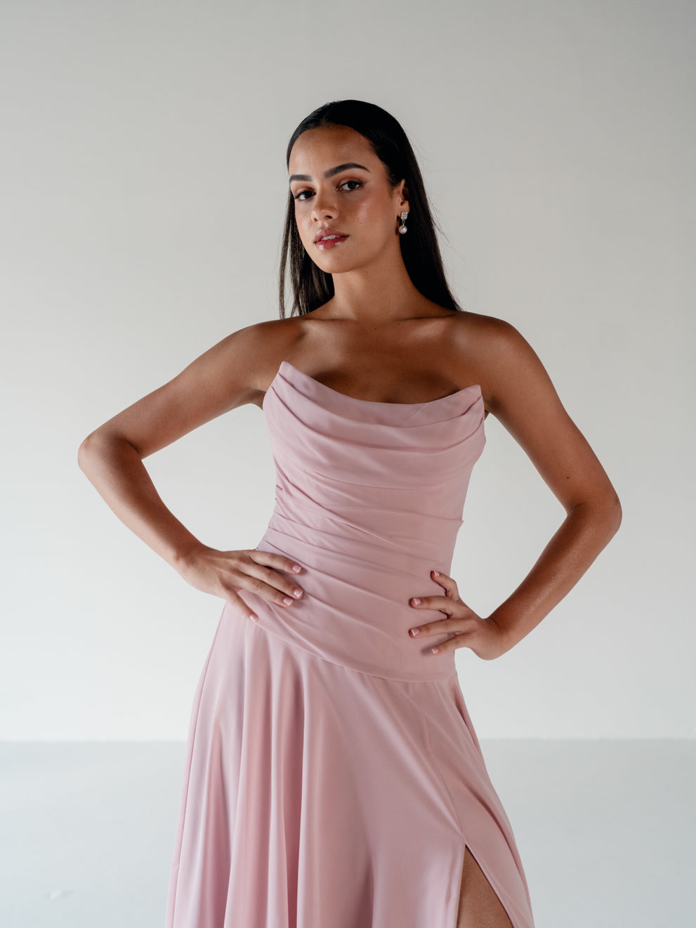 A woman modeling a strapless dusty pink dress with a draped, ruched bodice and a high side slit, set against a plain light gray studio background. / Strapless draped bodice with asymmetrical placed skirt and lace up back.