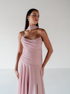 A woman modeling a dusty pink halter-neck midi dress with draped ruching on the bodice and a high slit, set against a plain light gray studio background. / Strapless draped bodice with asymmetrical placed skirt and lace up back.
