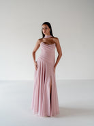 A woman modeling a floor-length dusty pink gown with a draped neckline, one-shoulder design, and a thigh-high slit, set against a plain light gray studio background. / Strapless draped bodice with asymmetrical placed skirt and lace up back.