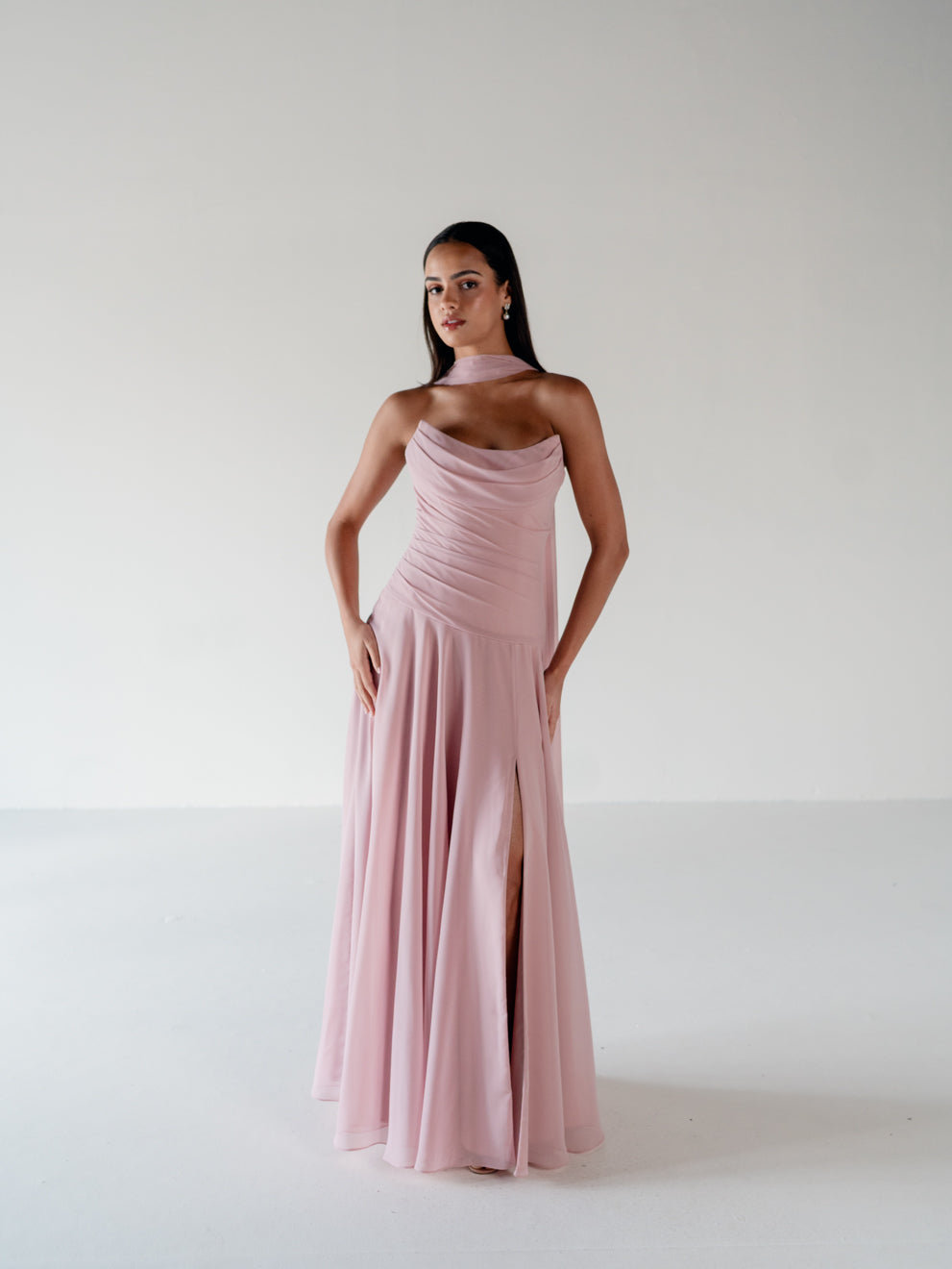 A woman modeling a floor-length dusty pink gown with a draped neckline, one-shoulder design, and a thigh-high slit, set against a plain light gray studio background. / Strapless draped bodice with asymmetrical placed skirt and lace up back.