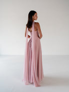 A woman stands with her back to the camera wearing a floor-length dusty pink gown with a draped shawl over one shoulder, photographed in a clean white studio setting. / Strapless draped bodice with asymmetrical placed skirt and lace up back.