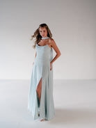 A woman modeling a floor-length light blue halter gown with a high thigh slit, photographed against a plain light gray studio background. / Strapless draped bodice with asymmetrical placed skirt and lace up back.