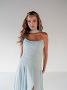 A woman modeling a light blue halter-neck midi dress with a draped, ruched bodice and a high leg slit, set against a plain light gray studio background. / Strapless draped bodice with asymmetrical placed skirt and lace up back.