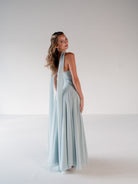 A woman modeling a floor-length light blue gown with a draped halter-style back, photographed in a clean studio setting against a light gray background. / Strapless draped bodice with asymmetrical placed skirt and lace up back.