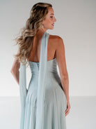 A woman modeling a light blue halter-neck maxi dress from the back, featuring a structured corset-style bodice and a long draped fabric detail, set against a plain light gray studio background. / Strapless draped bodice with asymmetrical placed skirt and lace up back.