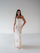 A woman modeling a strapless, floor-length floral gown in a bright, minimalist studio setting with a white background. / Scoop neckline fitted bodice with draped skirt.
