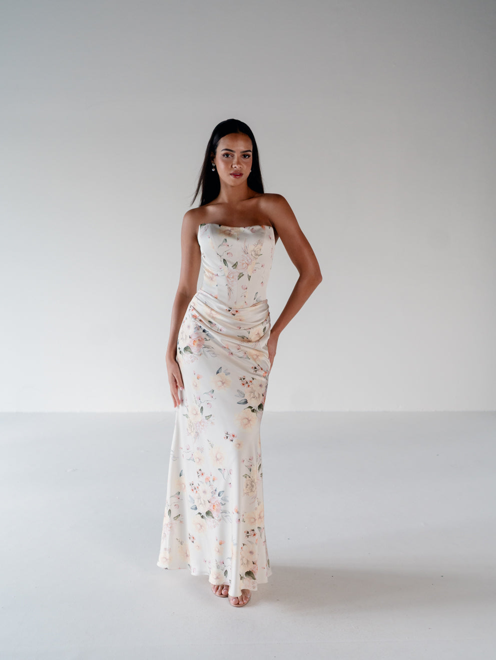 A woman modeling a strapless, floor-length floral gown in a bright, minimalist studio setting with a white background. / Scoop neckline fitted bodice with draped skirt.