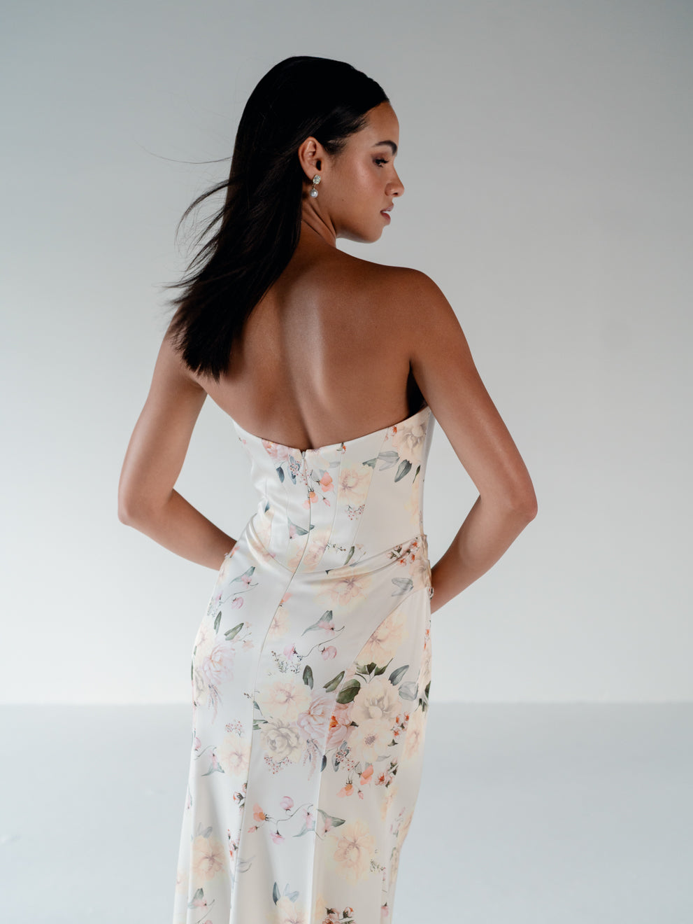 A woman shown from the back wearing a strapless fitted dress with a floral print in soft blush, peach, and green tones on a light cream base, against a plain light gray studio background. / Scoop neckline fitted bodice with draped skirt.