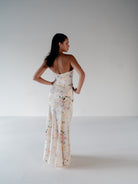 A woman stands with her back to the camera wearing a strapless, floor-length floral gown in a light cream color with pink, green, and blue floral details, set against a plain light gray studio background. / Scoop neckline fitted bodice with draped skirt.