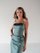A woman modeling a strapless satin midi dress in a dusty sage green color, standing against a neutral light gray studio background. / Scoop neckline fitted bodice with draped skirt.