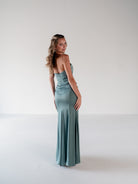 A woman modeling a strapless floor-length gown in a dusty sage green color, photographed from the back in a minimalist studio setting. / Scoop neckline fitted bodice with draped skirt.