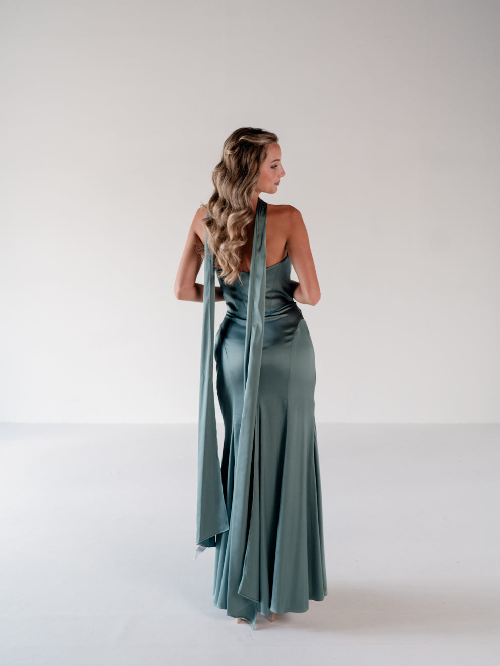 A woman stands with her back to the camera wearing a floor-length sage green satin gown with a draped halter-style back detail, photographed in a clean white studio setting. / Scoop neckline fitted bodice with draped skirt.