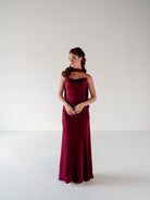 A woman modeling a floor-length burgundy evening gown with a halter-style neckline and draped ruffle detail, standing against a plain light gray studio background. / Scoop neckline fitted bodice with draped skirt.