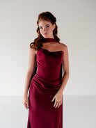A woman modeling a strapless, high-neck, floor-length gown in deep burgundy satin, photographed against a plain light gray studio background. / Scoop neckline fitted bodice with draped skirt.