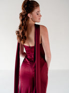 A woman shown from the back wearing a satin-like burgundy gown with a long draped veil or cape detail, photographed against a clean light gray studio background. / Scoop neckline fitted bodice with draped skirt.