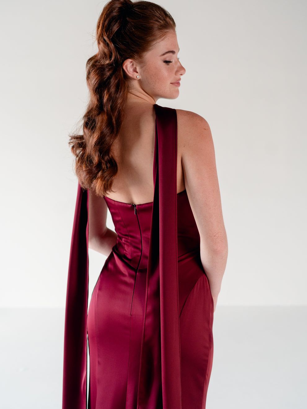 A woman shown from the back wearing a satin-like burgundy gown with a long draped veil or cape detail, photographed against a clean light gray studio background. / Scoop neckline fitted bodice with draped skirt.