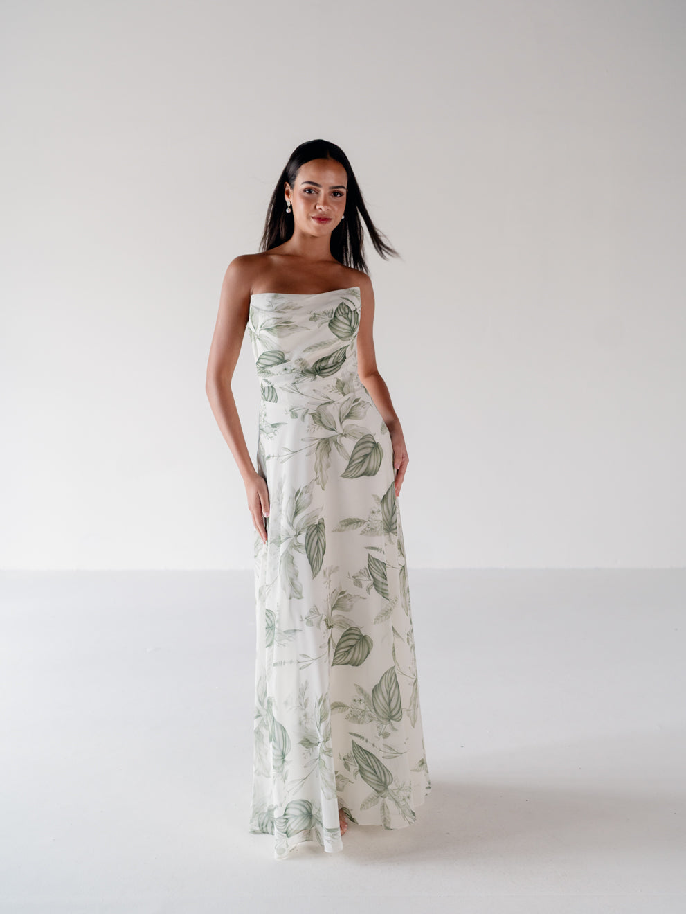A woman modeling a strapless maxi dress with a green botanical leaf print on a white background, standing in a minimalist studio setting. / Draped bodice with A-line skirt and lace up back.