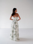 A woman stands with her back to the camera wearing a strapless white maxi dress with a green tropical leaf pattern, set against a plain white studio background. / Draped bodice with A-line skirt and lace up back.