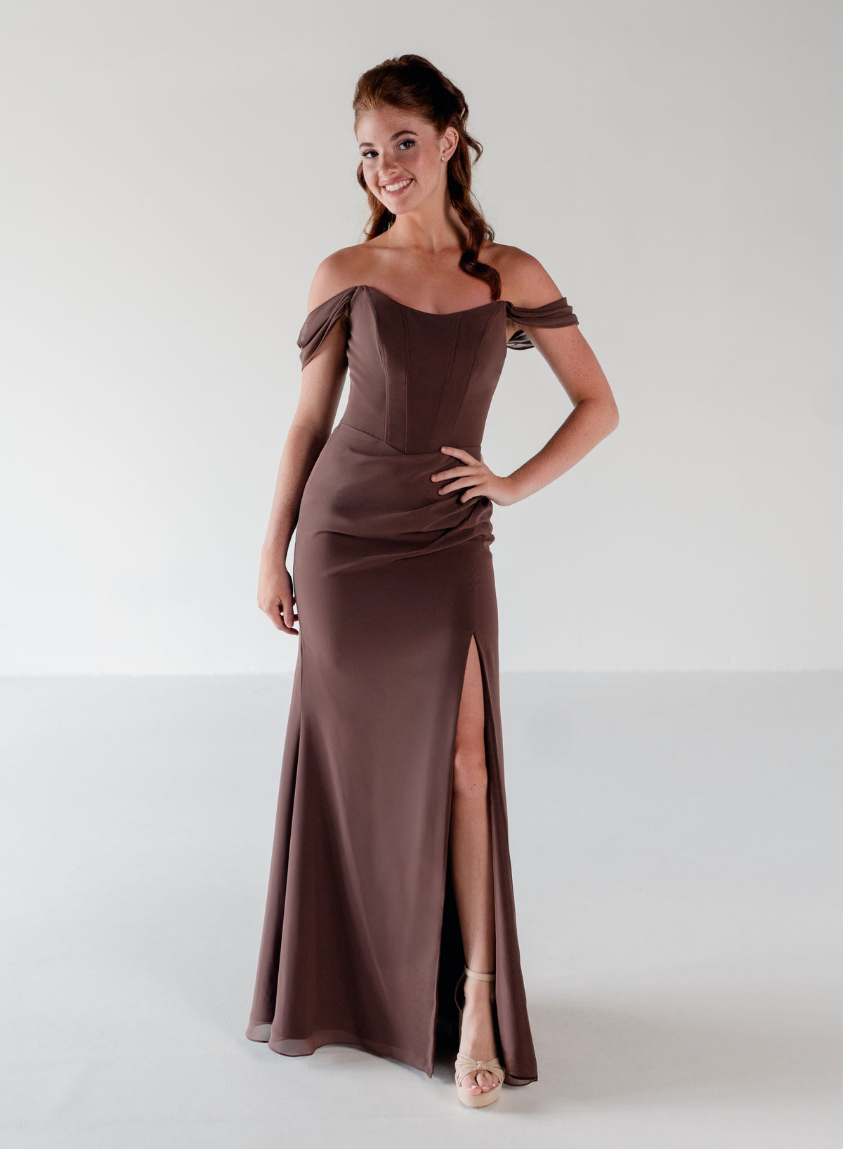 A woman modeling an off-the-shoulder floor-length gown in a muted taupe-brown color, featuring a fitted bodice, draped sleeves, and a high front slit, set against a plain light gray studio background. / Off the shoulder slim style with corset bodice detail and slit skirt.