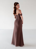 A woman modeling a floor-length off-the-shoulder gown in a muted taupe-brown shade, photographed against a clean light gray studio background. / Off the shoulder slim style with corset bodice detail and slit skirt.