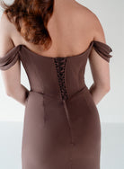 Rear view of a woman wearing an off-the-shoulder brown dress with a lace-up back corset detail, shown against a plain light background. / Off the shoulder slim style with corset bodice detail and slit skirt.