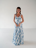 A woman modeling a sleeveless white maxi dress with a blue floral toile pattern, standing against a plain white studio background. / Draped neckline A-line gown with faux overskirt and slit detail.